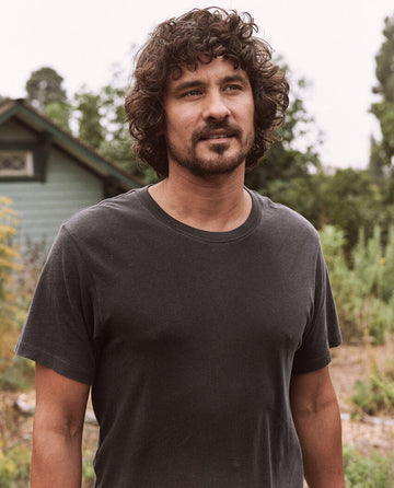 A man with curly hair and a beard is standing outside, wearing The Men's Slim Tee in Almost Black by THE GREAT. HOL 21 MENS, made from 100% cotton. In the background, there is greenery and part of a house.