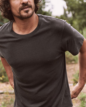 A person with a beard and mustache, wearing THE GREAT. HOL 21 MENS' The Men's Slim Tee in Almost Black, stands outdoors with greenery in the background. The image is cropped to show the upper torso and part of the face.