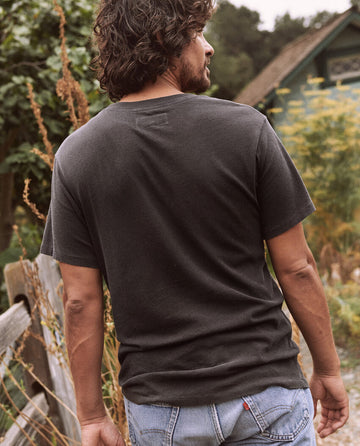 A man with shoulder-length hair, wearing The Men's Slim Tee in Almost Black by THE GREAT. HOL 21 MENS and jeans, is standing outdoors with his back turned, looking to his right. Trees and a house are visible in the background.