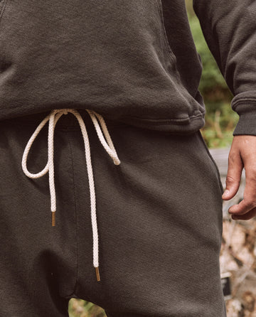 A close-up features a person wearing The Men's Stadium Sweatpant in Washed Black by THE GREAT. HOL 21 MENS, complemented by a matching dark hoodie. The sweatpants have a white drawstring, giving off a vintage athleticwear vibe. One hand is partially visible on the right side of the image, embodying the casual Los Angeles street style.
