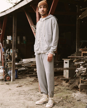 A person in The Men’s Zip Up Hoodie -- Varsity Grey from THE GREAT. SP22 D2 MENS and sweatpants stands outside an old wooden shed. They are wearing a brown beanie and white sneakers. Various tools and materials are scattered around the shed, highlighting the versatile nature of their attire.