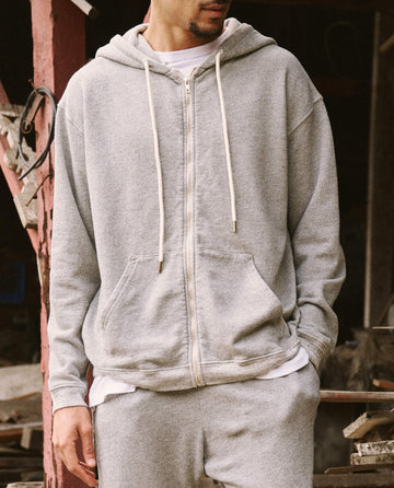 A person wearing a light grey The Men's Zip Up Hoodie by THE GREAT. SP22 D2 MENS, in the Varsity Grey color, and matching sweatpants made from ultra-soft cotton stands with hands in pockets in an outdoor setting. The versatile outfit perfectly blends comfort and style.