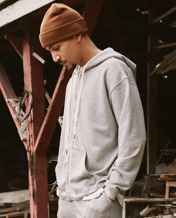 A person wearing The Men's Zip Up Hoodie in Varsity Grey by THE GREAT. SP22 D2 MENS and a brown beanie stands with hands in pockets, looking downward outdoors near a wooden structure. The versatile hoodie, made from ultra-soft cotton, adds comfort to the scene.