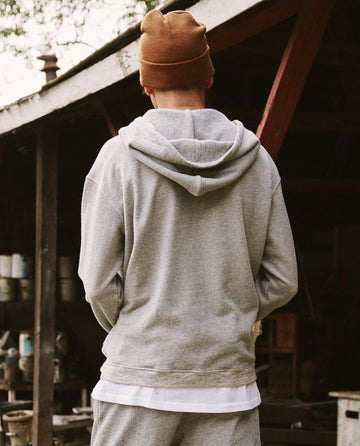 Person in a versatile Varsity Grey Zip Up Hoodie from THE GREAT. SP22 D2 MENS and a brown beanie, facing away, standing near a rustic wooden structure with shelves and tools.