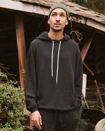 A person wearing The Men's Gym Drawstring Hoodie in Almost Black by THE GREAT. SU22 MENS, along with a green beanie, stands outdoors in front of a rustic wooden building with plants nearby.