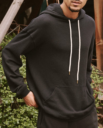 A person stands outdoors wearing The Men's Gym Drawstring Hoodie in Almost Black from THE GREAT. SU22 MENS, which features white drawstrings and is made of softly washed cotton with a traditional front kangaroo pocket. One hand rests comfortably in their pocket, with green foliage visible in the background.