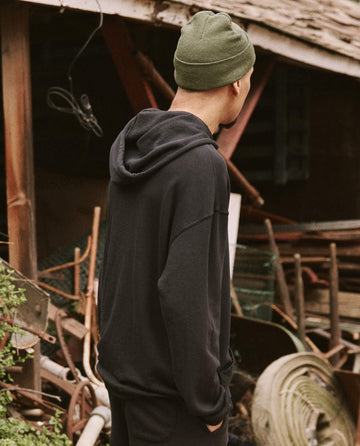A person wearing THE GREAT. SU22 MENS Men's Gym Drawstring Hoodie in Almost Black and a green beanie stands outdoors, facing away, with a rustic background of wooden structures and various outdoor equipment.