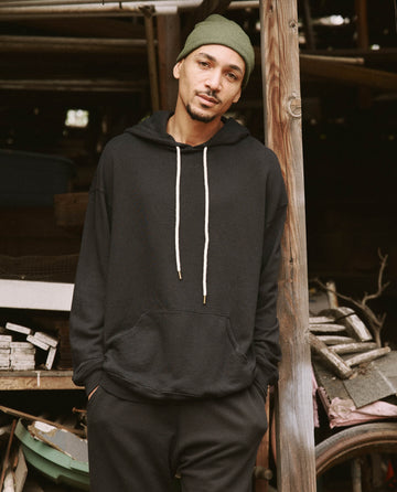 A man wearing THE GREAT. SU22 MENS "The Men's Gym Drawstring Hoodie" in Almost Black and a green beanie stands casually next to a wooden post, with various stacked items in the background. The softly washed cotton of his vintage athleticwear adds to his relaxed vibe.
