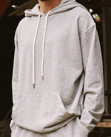 A person wearing THE GREAT. SU22 MEN'S The Men's Gym Drawstring Hoodie in Varsity Grey stands outdoors. The face is not visible.