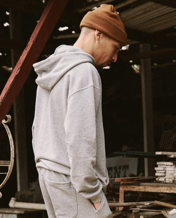 A person in THE GREAT. SU22 MENS's The Men's Gym Drawstring Hoodie in Varsity Grey and a brown beanie stands outdoors, looking downward with hands in pockets.