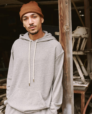 A man wearing The Men's Gym Drawstring Hoodie in Varsity Grey from THE GREAT. SU22 MENS and a brown beanie, both made in the U.S.A., stands next to a wooden post in an outdoor setting.
