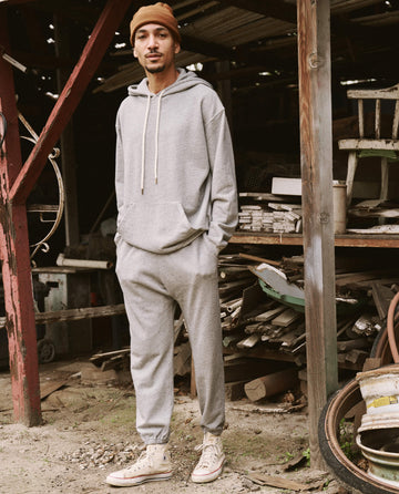A man wearing THE GREAT. SU22 MENS' Varsity Grey Men's Gym Drawstring Hoodie, matching sweatpants, and white sneakers stands in a cluttered outdoor area with various wooden and metal objects around him.