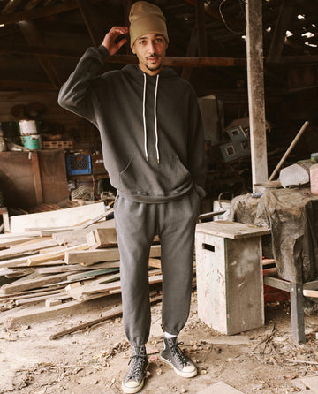 A man in a dark hoodie and The Men's Stadium Sweatpant by THE GREAT. HOL 21 MENS in Washed Black, wearing a beige beanie and sneakers, stands in an old workshop surrounded by wooden planks and tools, evoking a sense of timeless craftsmanship.