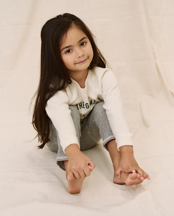 A young girl with long dark hair sits on the floor, barefoot and smiling slightly, wearing The Little College Sweatshirt in washed white with a hornet graphic from THE GREAT. SP22 D2 LITTLE, paired with gray pants.