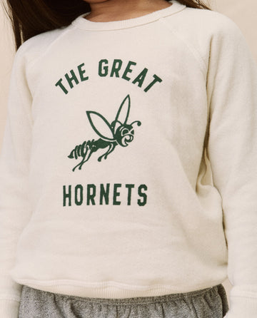 A child wearing The Little College Sweatshirt from THE GREAT. SP22 D2 LITTLE, featuring a washed white, 100% cotton design with green text that reads "The Great Hornets" and an illustration of a hornet in the center, giving it a charming vintage wash.