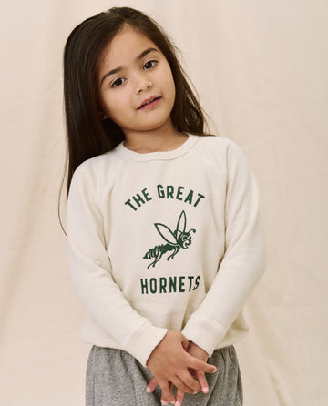 A young child with long hair, wearing THE GREAT. SP22 D2 LITTLE's "The Little College Sweatshirt" in washed white featuring a hornet graphic and "The Great Hornets" text, stands with hands clasped in front.