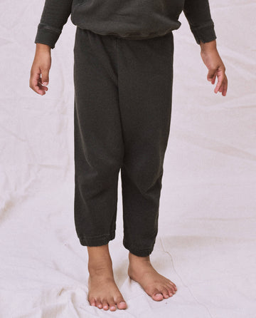 A child is wearing The Little Stadium Sweatpant in Washed Black from THE GREAT. SP22 D2 LITTLE, made of 100% cotton and inspired by vintage athletic attire. They are standing barefoot on a light-colored surface, with their upper body partially cropped out of the frame.