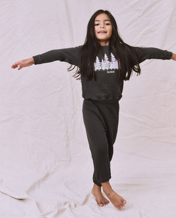 A young girl with long hair is standing with arms outstretched, wearing The Little Stadium Sweatpant in Washed Black featuring a tree design on the front that echoes vintage athletic wear from THE GREAT. SP22 D2 LITTLE. The background is a light-colored fabric.