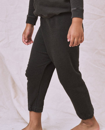 A person standing barefoot against a plain background, wearing vintage athletic wear in the form of The Little Stadium Sweatpant in Washed Black by THE GREAT. SP22 D2 LITTLE. The image is cropped to show only the lower half of their body.