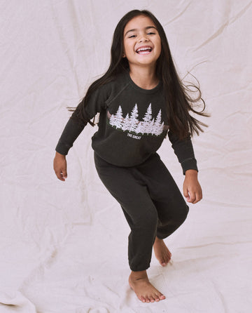 A young girl with long dark hair is standing on one leg, smiling. She is dressed in a black long-sleeve shirt featuring a white tree design and The Little Stadium Sweatpant in Washed Black from THE GREAT. SP22 D2 LITTLE, both made of 100% cotton. The background appears to be a light fabric.