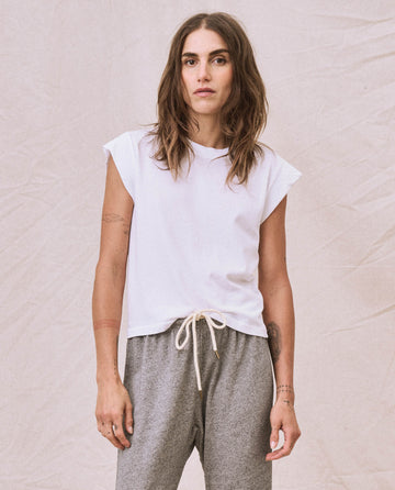 A person with long hair stands against a fabric backdrop, wearing The Peak Shoulder Tee in True White from THE GREAT. FALL 22 KNITS and grey drawstring pants.