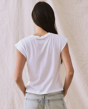 Person with long dark hair wearing a True White "The Peak Shoulder Tee" by THE GREAT. FALL 22 KNITS and light blue jeans, facing away, against a light-colored draped fabric background.