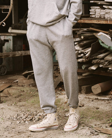 A person standing on a dirt ground, scattered with wooden planks and metal pieces in the background, is wearing The Men's Stadium Sweatpant - Varsity Grey by THE GREAT. HOL 21 MENS and white high-top sneakers.