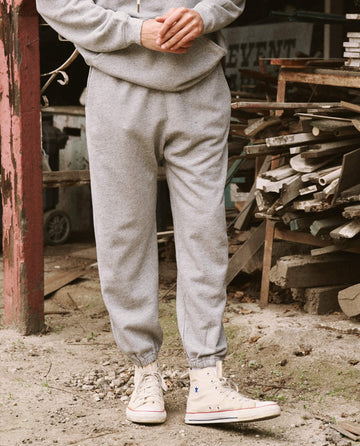 A person wearing THE GREAT. HOL 21 MENS' The Men's Stadium Sweatpant in Varsity Grey and high-top sneakers stands in a rustic setting with scattered lumber and old furniture in the background.