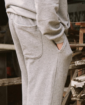 Close-up of a person wearing THE GREAT. HOL 21 MENS The Men's Stadium Sweatpant in Varsity Grey, with their hand in the pocket. The background features blurred wooden structures and tools, suggesting a workshop or garage setting.