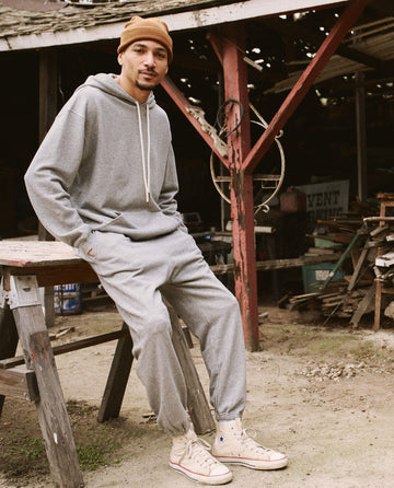 A person in a gray hoodie, The Men's Stadium Sweatpant by THE GREAT. HOL 21 MENS in Varsity Grey, and white sneakers, wearing a brown beanie, leans against a weathered wooden table in an outdoor setting with various tools and objects in the background.