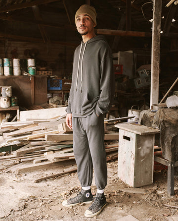 A person stands in a Los Angeles workshop wearing a beanie, hoodie, and vintage athleticwear including The Men's Stadium Sweatpant in Washed Black by THE GREAT. HOL 21 MENS, with hands in pockets. The workshop has scattered wooden planks, paint cans, and various tools.