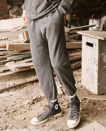A person standing in a workshop is wearing THE GREAT. HOL 21 MENS' The Men's Stadium Sweatpant in Washed Black, paired with black high-top sneakers and white socks. The background includes wooden planks and a green workbench, creating a scene that exudes vintage athleticwear vibes, perfect for the laid-back lifestyle of Los Angeles.