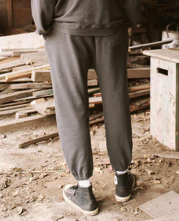 A person wearing The Men's Stadium Sweatpant in Washed Black from THE GREAT. HOL 21 MENS and black high-top sneakers stands with their back to the camera in a cluttered workshop with wooden boards on the floor, exuding a vintage athleticwear vibe reminiscent of Los Angeles street style.