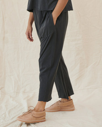 A person wearing dark pants and light brown moccasin-style shoes stands with one hand in their pocket against a neutral backdrop, showcasing *The Jersey Crop. - Washed Black* by THE GREAT. CORE KNITS, made from 100% Cotton fabric.