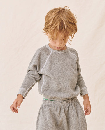 A young child with blond hair, wearing THE GREAT. FALL21 D2 LITTLE's The Little College Sweatshirt in Varsity Grey and soft polyester and cotton sweatpants, stands against a neutral background looking down.
