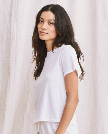 A person with long dark hair wearing "The Little Tee" in True White by THE GREAT. SP22 APRILCAP, reminiscent of a vintage 1930s undershirt, stands against a light-colored fabric backdrop.