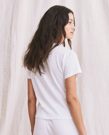 A person with long, dark hair is standing and facing slightly to the right, wearing The Little Tee in True White from the brand THE GREAT. SP22 APRILCAP, which looks reminiscent of a vintage 1930s undershirt, in front of a light-colored curtain.