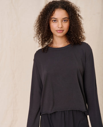 A woman with curly hair is showcasing The Long Sleeve Crop Tee in Almost Black by THE GREAT. CORE KNITS, standing against a plain beige background.