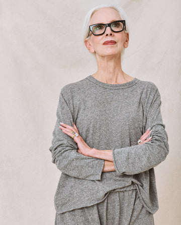 An elderly woman with short white hair and black glasses stands with her arms crossed. She wears *The Long Sleeve Crop Tee in Heather Grey* from THE GREAT. CORE KNITS, layered over a square cropped tee and matching pants, looking upward with a neutral expression.