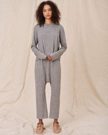 A person with curly hair stands against a neutral background, wearing a loose-fitting gray jumpsuit layered over THE GREAT. CORE KNITS' The Long Sleeve Crop Tee in Heather Grey and beige shoes.