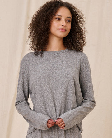 A woman with curly hair, wearing The Long Sleeve Crop Tee in Heather Grey from THE GREAT. CORE KNITS, stands against a plain beige background.