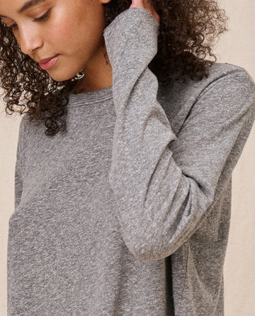 Person in "The Long Sleeve Crop Tee" in heather grey by THE GREAT. CORE KNITS, with curly hair and a calm expression, layers effortlessly with a square cropped tee featuring a raw hem.