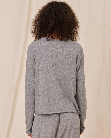 Person with curly hair wearing The Long Sleeve Crop Tee in Heather Grey by THE GREAT. CORE KNITS, paired with matching pants, shown from the back. The background is a plain, light-colored curtain or wall.