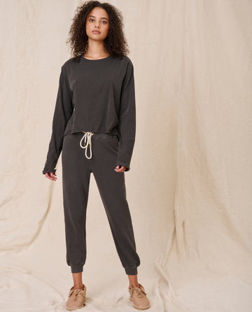 A person stands against a beige fabric background, wearing a loose-fitting long sleeve crop tee in washed black from THE GREAT. CORE KNITS and matching pants with a white drawstring. They finish the look with light brown shoes, and the ensemble is completed with a raw hem that adds an edgy touch.