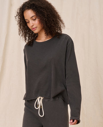 A person in casual dark clothing with curly hair stands against a beige background, wearing The Long Sleeve Crop Tee in Washed Black from THE GREAT. CORE KNITS, looking slightly downward.