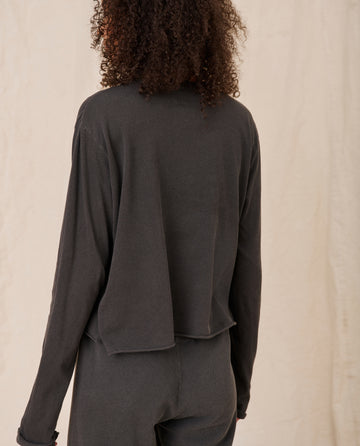 Person with curly hair facing away, wearing The Long Sleeve Crop Tee in Washed Black from THE GREAT. CORE KNITS and black pants, standing against a light-colored background.