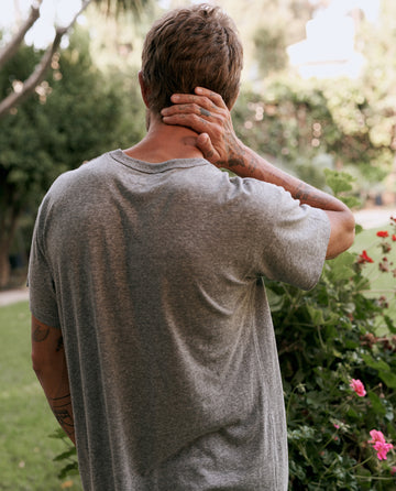 A person with short hair wearing The Men's Boxy Crew in Heather Grey by THE GREAT. MAN stands outdoors, facing away, holding the back of their neck with one hand. A garden with green shrubs and blooming flowers is in the background.