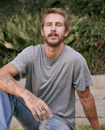 A person with tattoos on both arms, wearing The Men's Boxy Crew in Heather Grey from THE GREAT. MAN and blue jeans made in the U.S.A., is sitting outdoors on a stone path with greenery in the background.