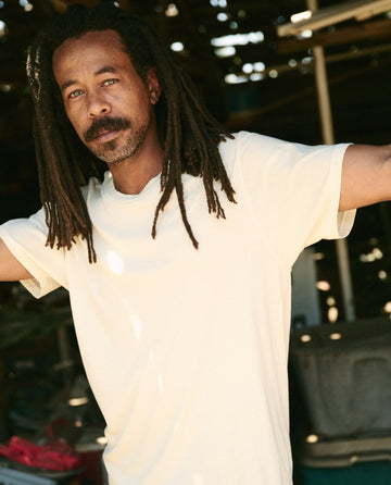 A man with long dreadlocks and a beard, wearing The Men's Boxy Crew in washed white by THE GREAT. MAN, stands with his arms slightly raised in an outdoor setting.