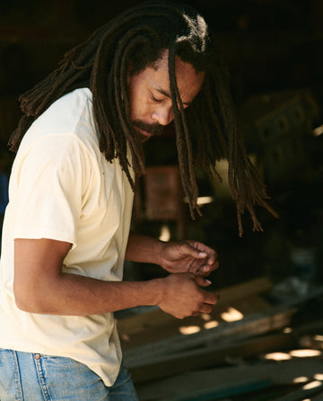 A person with long dreadlocks wearing a perfectly fitting washed white cotton tee from The Men's Boxy Crew by THE GREAT. MAN and jeans is looking down while working with their hands in an indoor setting.